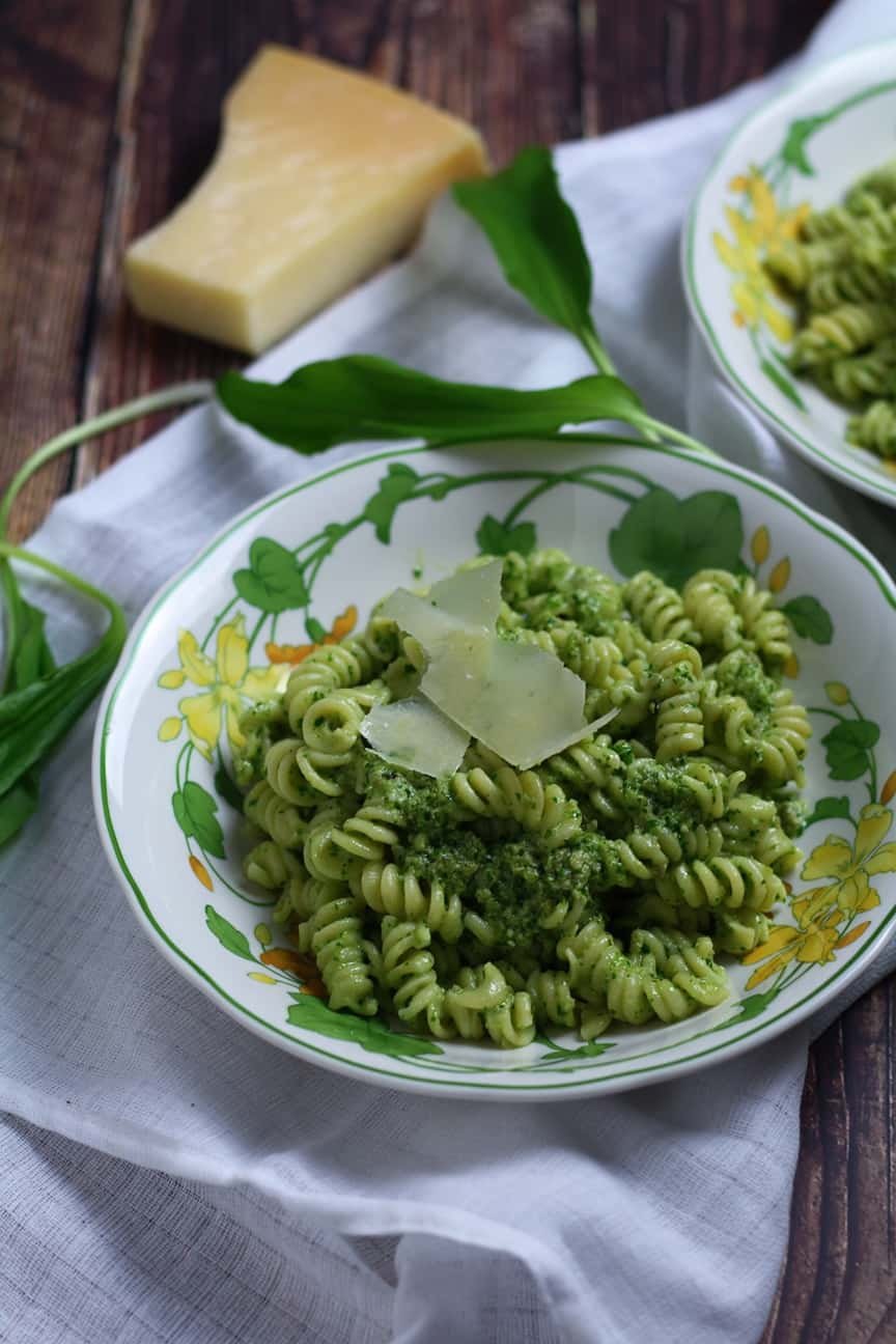 Two bowls of Wild Garlic Pesto Pasta on a table with wild garlic leaves and a block of Parmesan cheese.