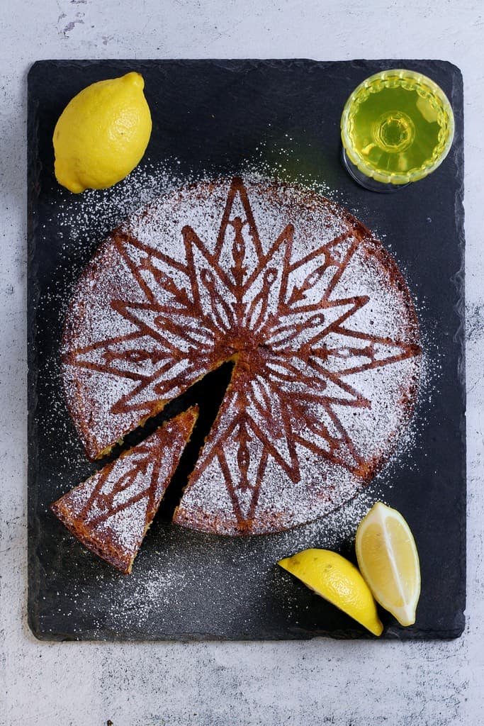 Torta Caprese al limone or Capri Lemon cake on a slate board with lemons, a glass of limoncello and a cake server.