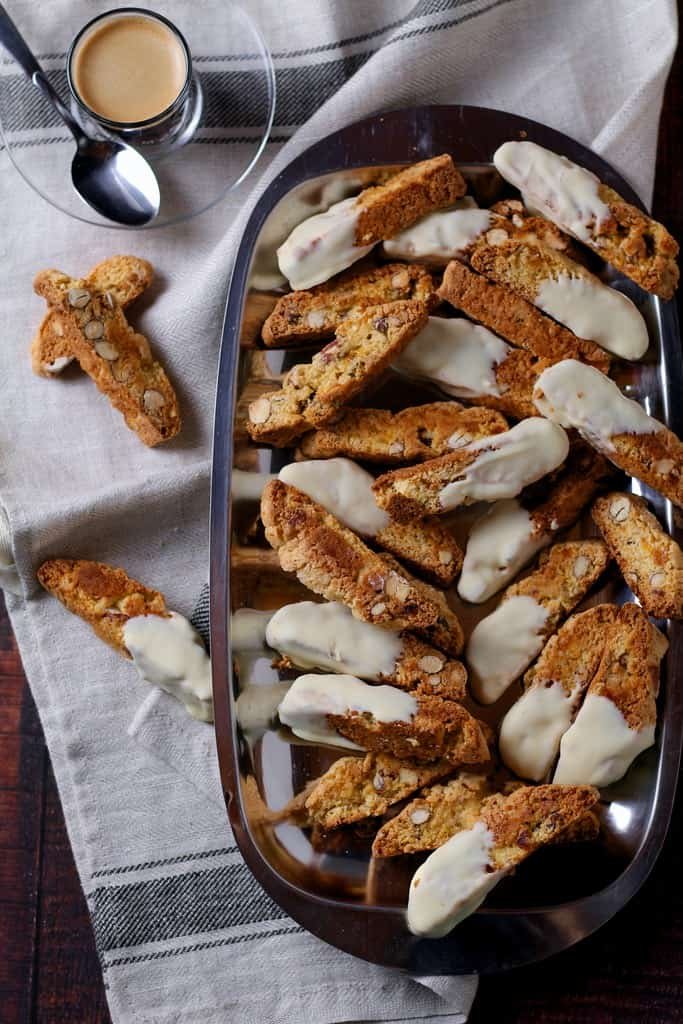 A tray of white chocolate dipped, apricot and almond biscotti on a tray with a small glass of espresso in the background.