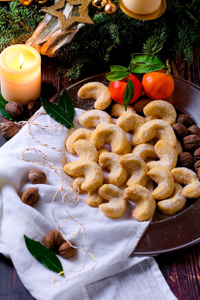 A plate of Vanillekipferl or Vanilla crescent cookies surrounded by Christmas decorations, walnuts, clementines and a candle.