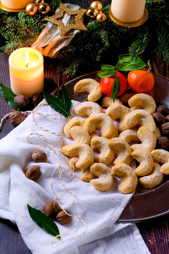 A plate of Vanillekipferl or Vanilla crescent cookies surrounded by Christmas decorations, walnuts, clementines and a candle.