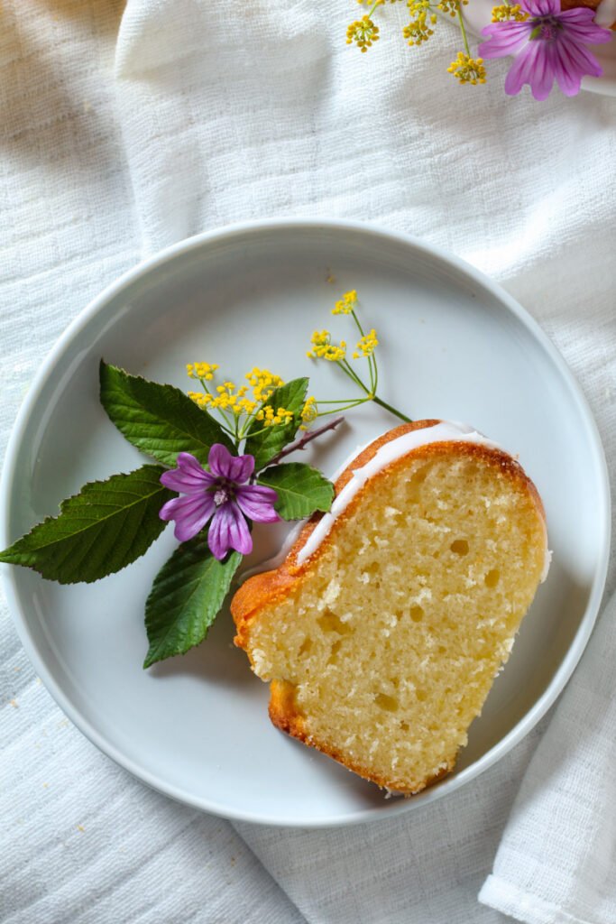 A slice of lemon bundt cake on a plate with fresh mallow and fennel flowers.