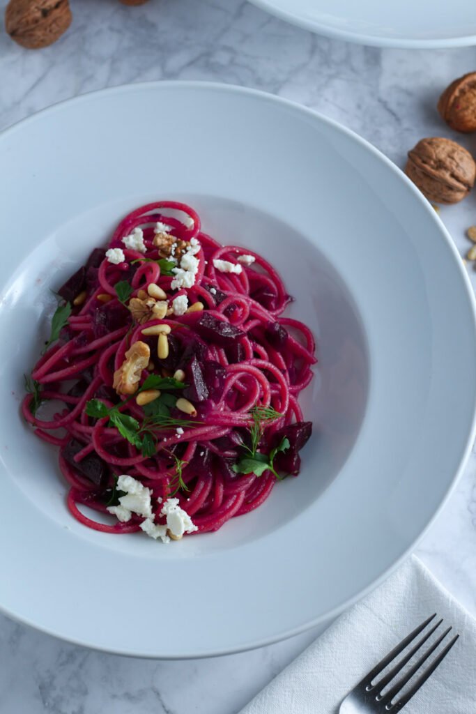 A picture of Beetroot and Feta Pasta in a white bowl, topped with toasted nuts and feta cheese