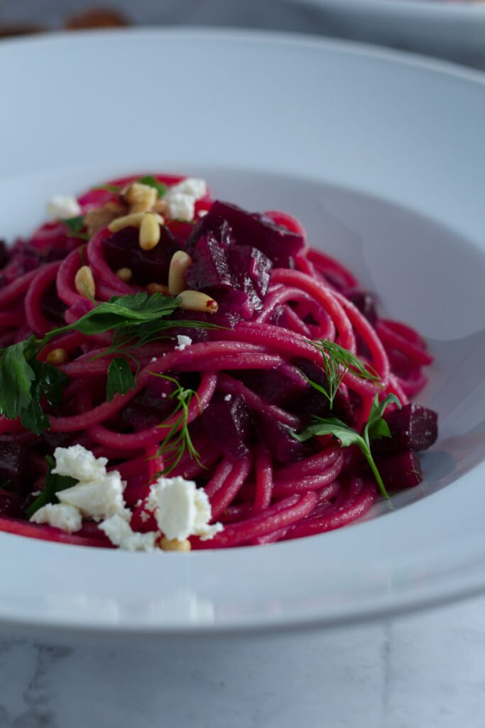 A picture of Beetroot and Feta Pasta in a white bowl, topped with toasted nuts and feta cheese