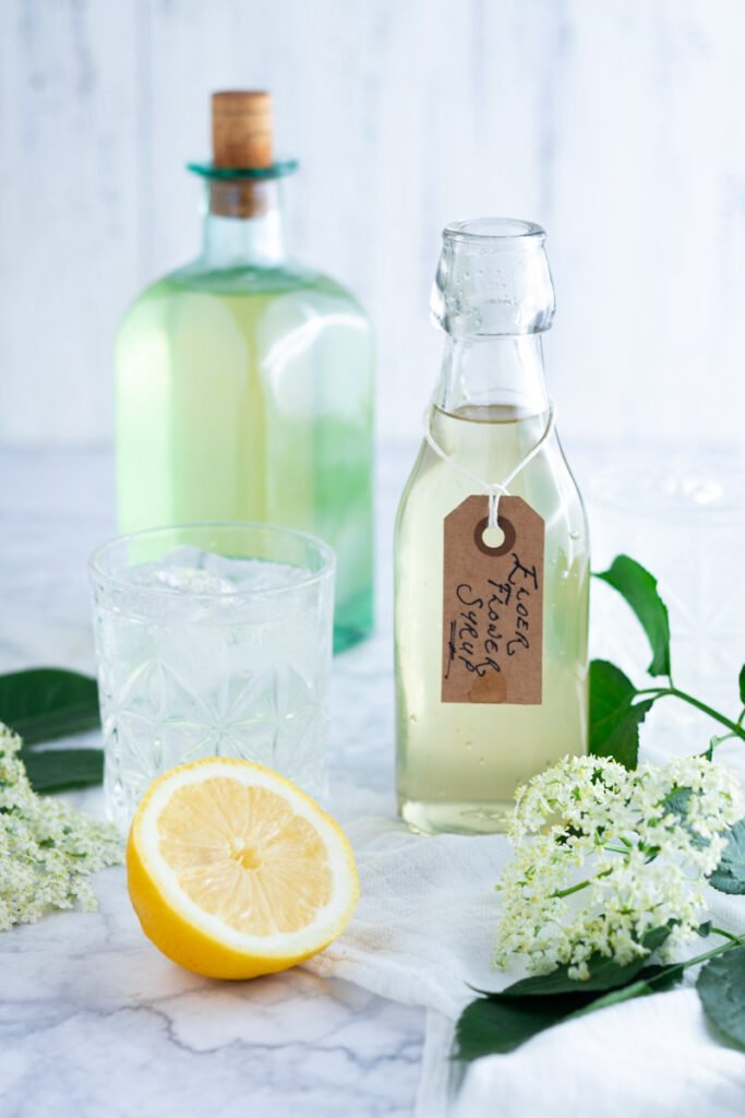 A bottle of elderflower cordial (syrup) with fresh elderflowers, and lemons.