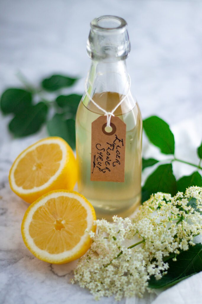 A bottle of elderflower cordial (syrup) with fresh elderflowers, and lemons.
