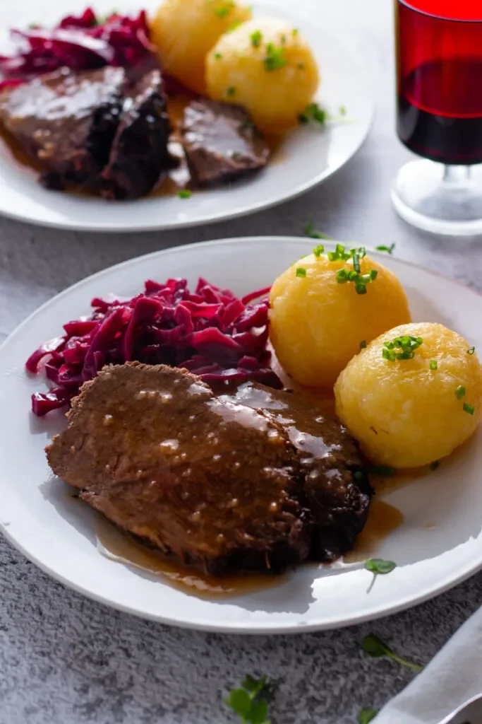 German Sauerbraten with red cabbage and potato dumplings on a plate with sauce and a glass of red wine.
