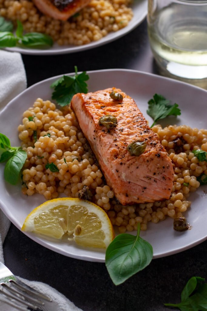 Lemon and Caper Salmon with herby couscous on a serving plate with a glass of white wine.