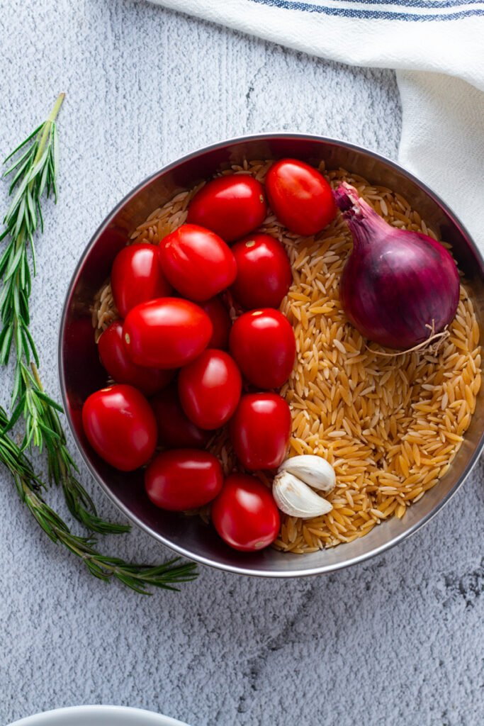 Rosemary, cherry tomatoes, orzo, garlic and red onions in a bowl.