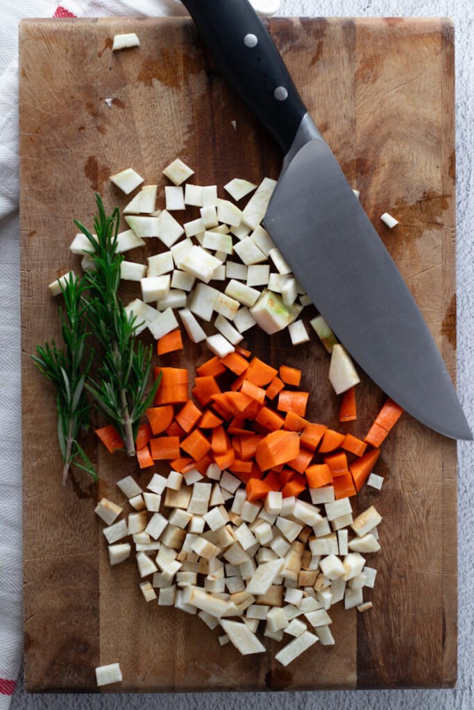 Diced parsnip, celeriac and carrot with a chef's knife on a wooden chopping board.