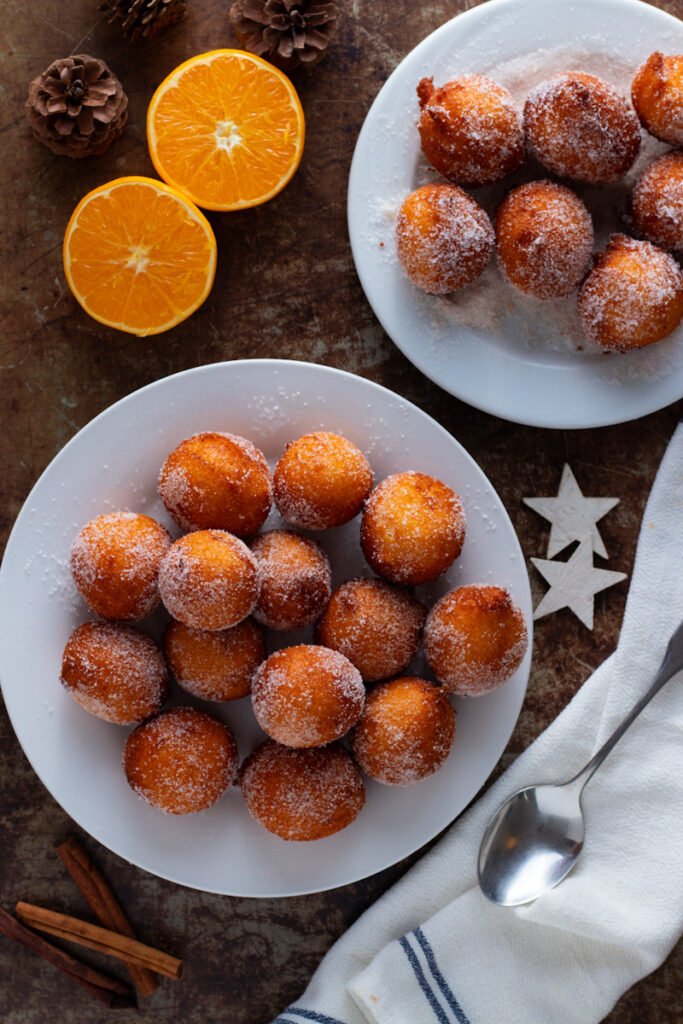Plates of Quarkbällchen donut holes on a table with fresh oranges.