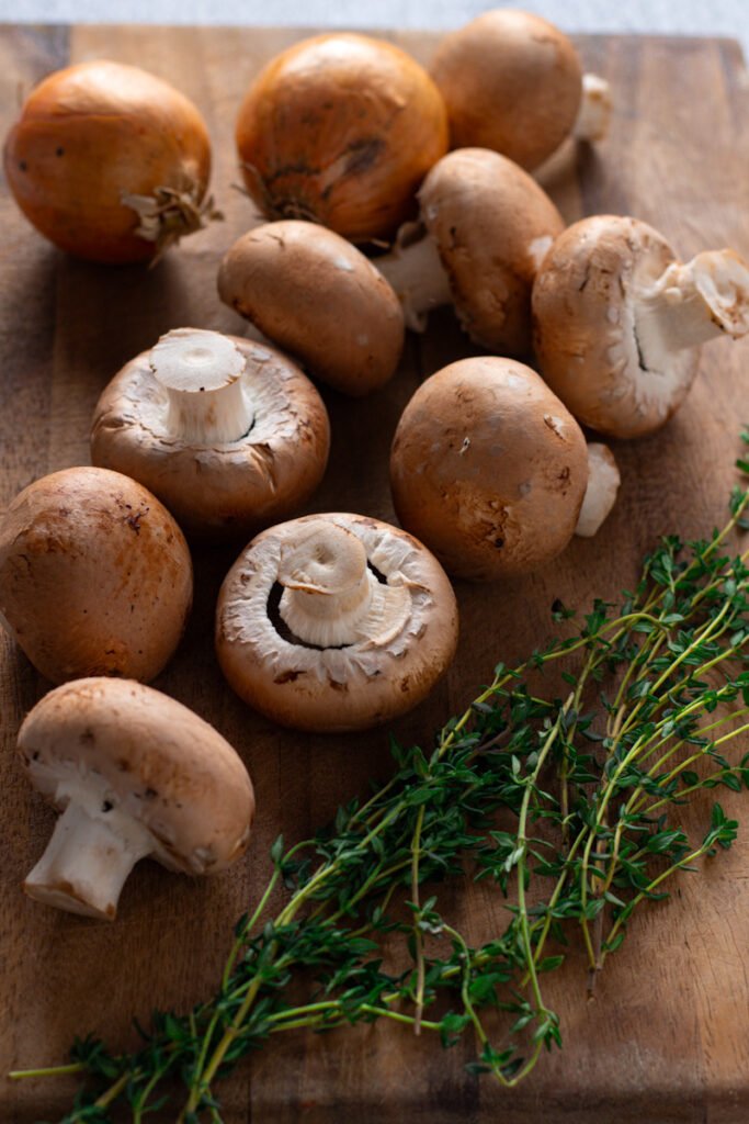 Brown mushrooms, onions and fresh thyme on a chopping board.