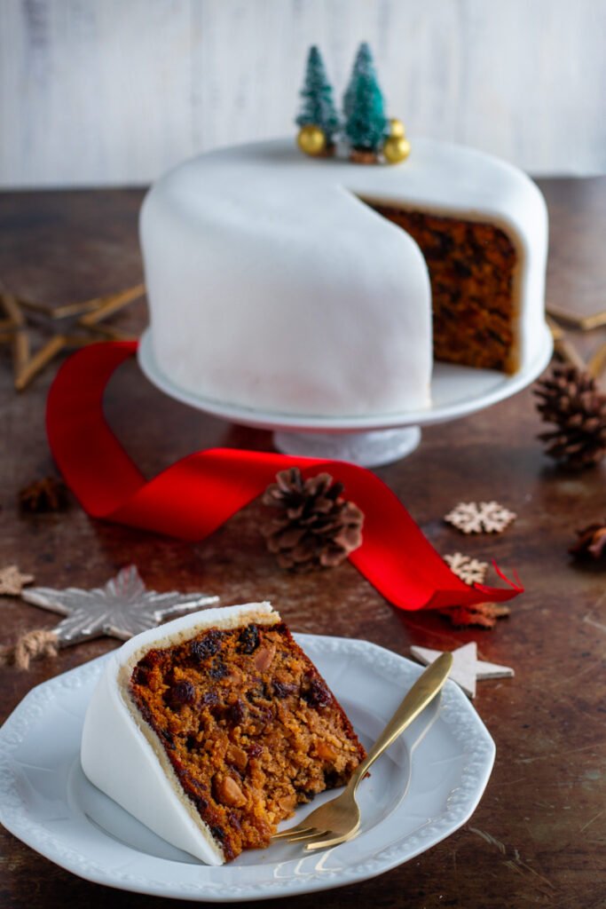 A slice of Christmas cake with the whole Christmas Cake in the background.