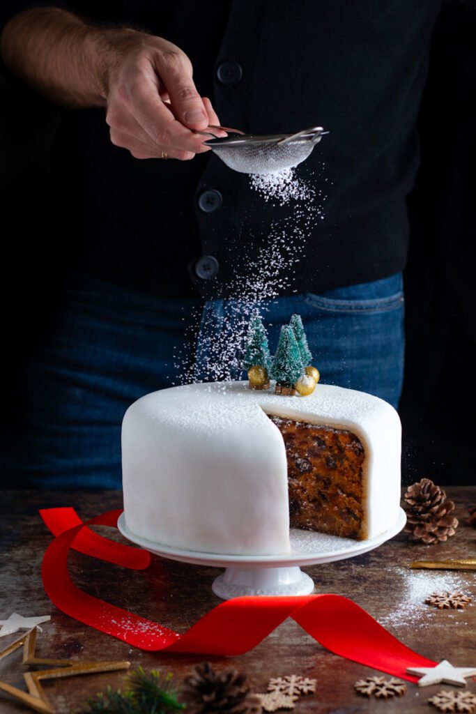 A traditional English Christmas cake on a cake stand being sprinkled with powdered sugar by a man standing behind.