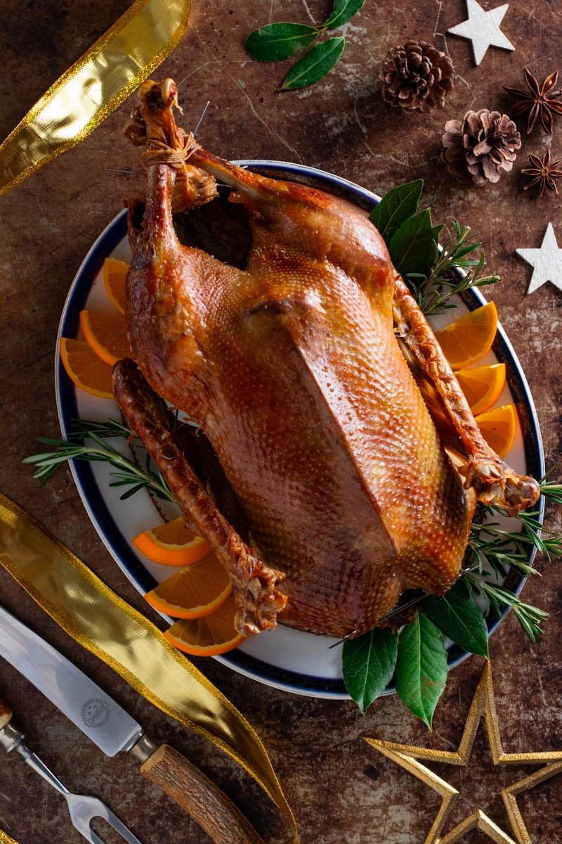 A roast goose seen from above on a table with herbs, oranges, and carving utensils.