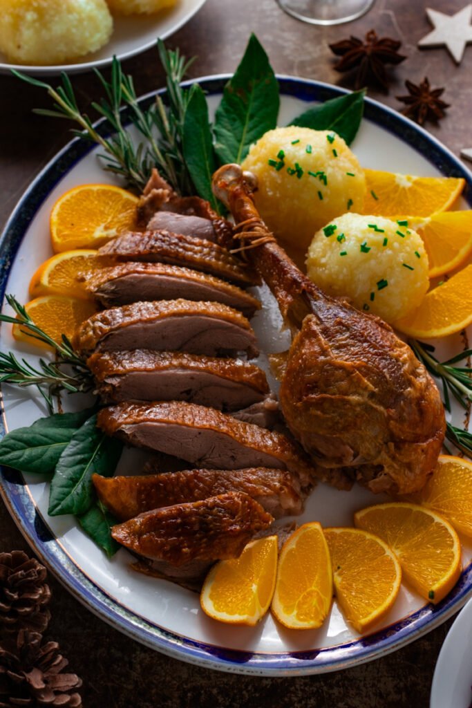 A plate of sliced roast goose with potato dumplings, orange slices and fresh herbs.