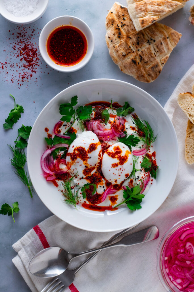 Turkish Poached Eggs in a bowl on a table with fresh herbs, red pepper oil and bread.