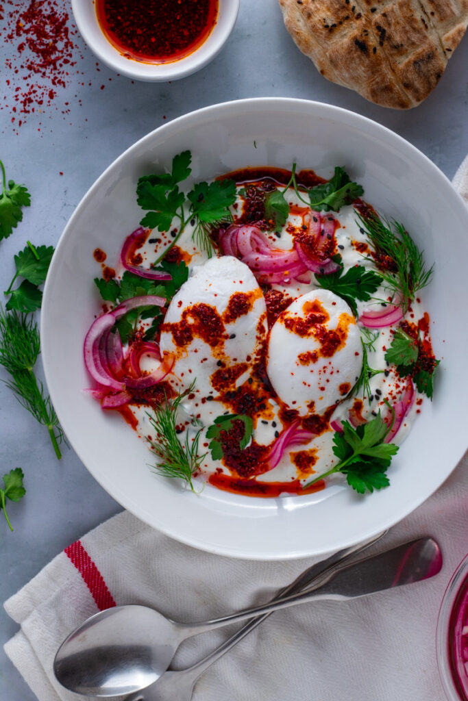 Turkish Poached Eggs in a bowl on a table with fresh herbs, red pepper oil and bread.
